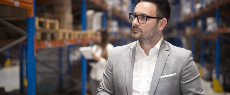 Portrait of successful businessman manager CEO holding tablet and walking through warehouse storage area looking towards shelves.
